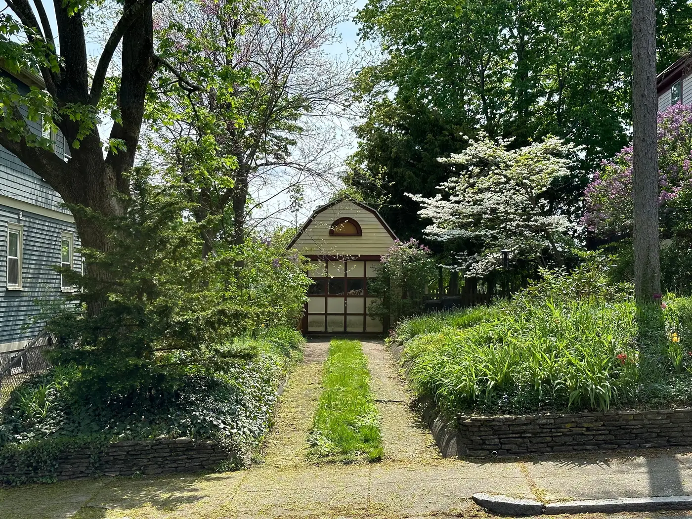 Detached gambrel-roof shed behind a house, reached by a grass path through a garden